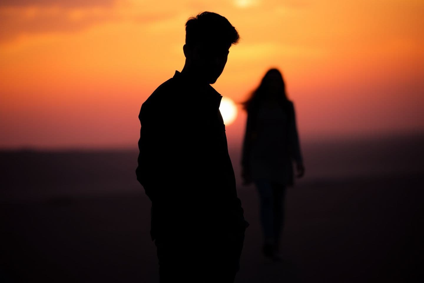 AI generated: A cinematic emotional scene of a young man standing alone during sunset, orange and purple sky, soft sunlight, silhouette style, a girl walking away in the distance slightly blurre